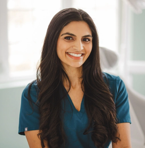 Professional woman with long dark hair smiling in dental office setting wearing blue scrubs.