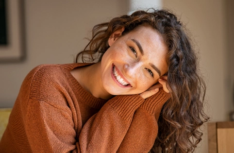 Woman smiling warmly indoors wearing rust-colored sweater in comfortable home setting