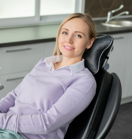 Woman in light purple shirt sitting in dental chair in modern office setting, smiling at camera.