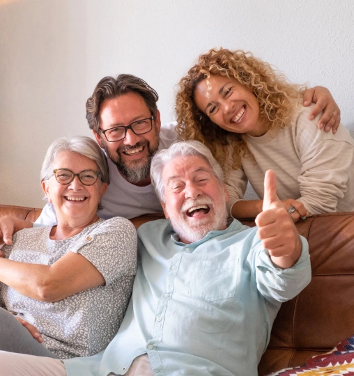Four happy multigenerational family members smiling together indoors, showing thumbs up gesture.