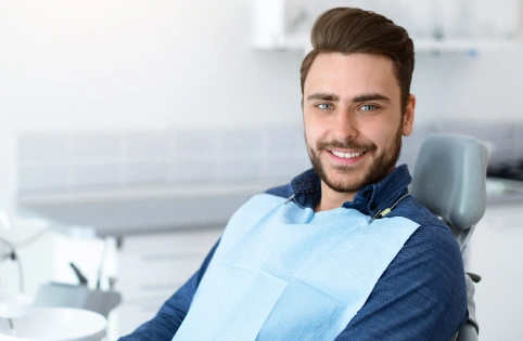 Smiling male dentist in blue scrubs and jacket sitting in modern dental office setting.