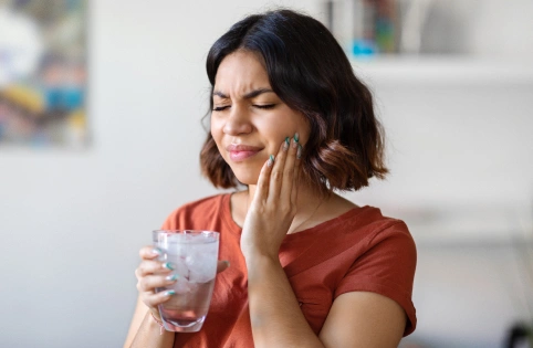 Woman holding glass of water touching her cheek in pain, suggesting tooth sensitivity or dental discomfort.