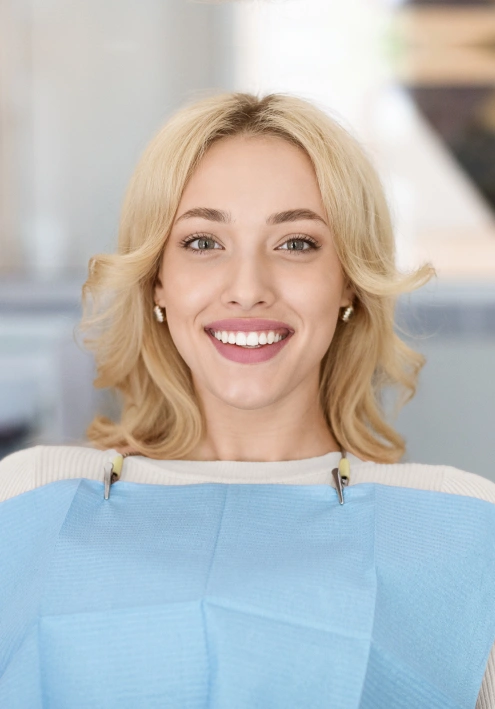 Woman with blonde wavy hair smiling at camera in light blue top, professional dental office setting.