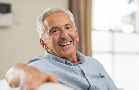 Smiling older man in light blue shirt gesturing welcomingly in bright dental office setting.