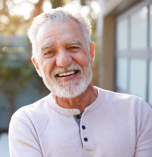 Smiling older man with white hair and beard wearing light purple shirt outdoors.