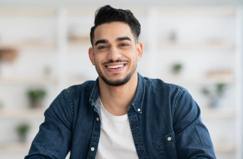 Smiling man in blue shirt and white t-shirt in bright, blurred office setting.